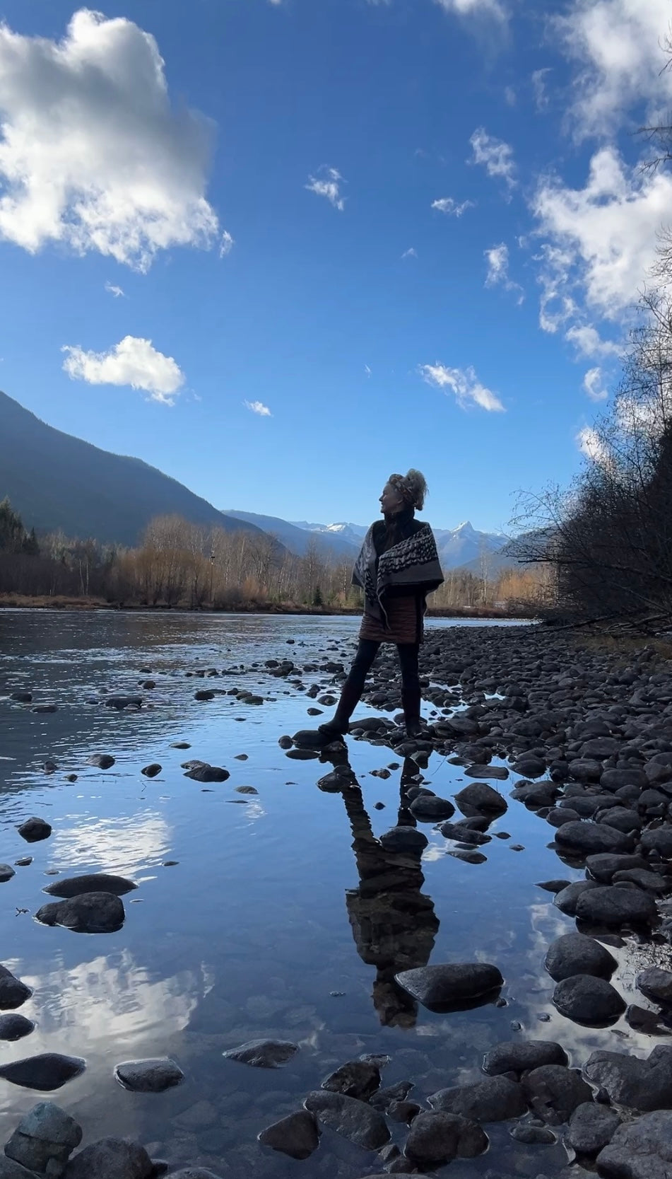 Irene Rutherford stands at Slocan River'sedge in an owl shawl with Frog Peak behind her as she looks to the East